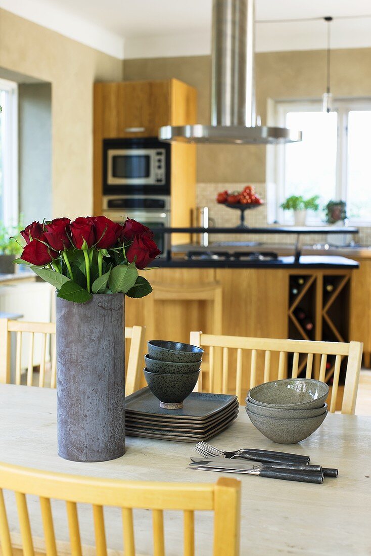 A grey vase or red roses, grey plates and bowls on a table with a view of a kitchen counter