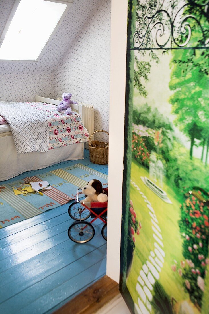A child's bedroom with a blue-painted wooden floor, a bed under a skylight window and a door decorated with a photo of a garden