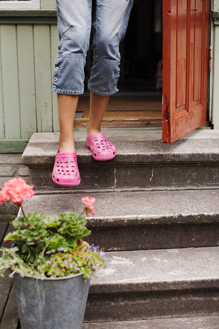 A child wearing pink shoes standing by a front door with a potted plant at the bottom of a three stone steps