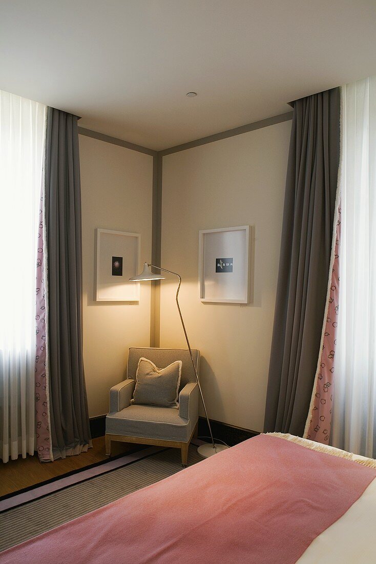 Light gray armchair with a floor lamp in the corner of a bedroom and floor length curtains at the window