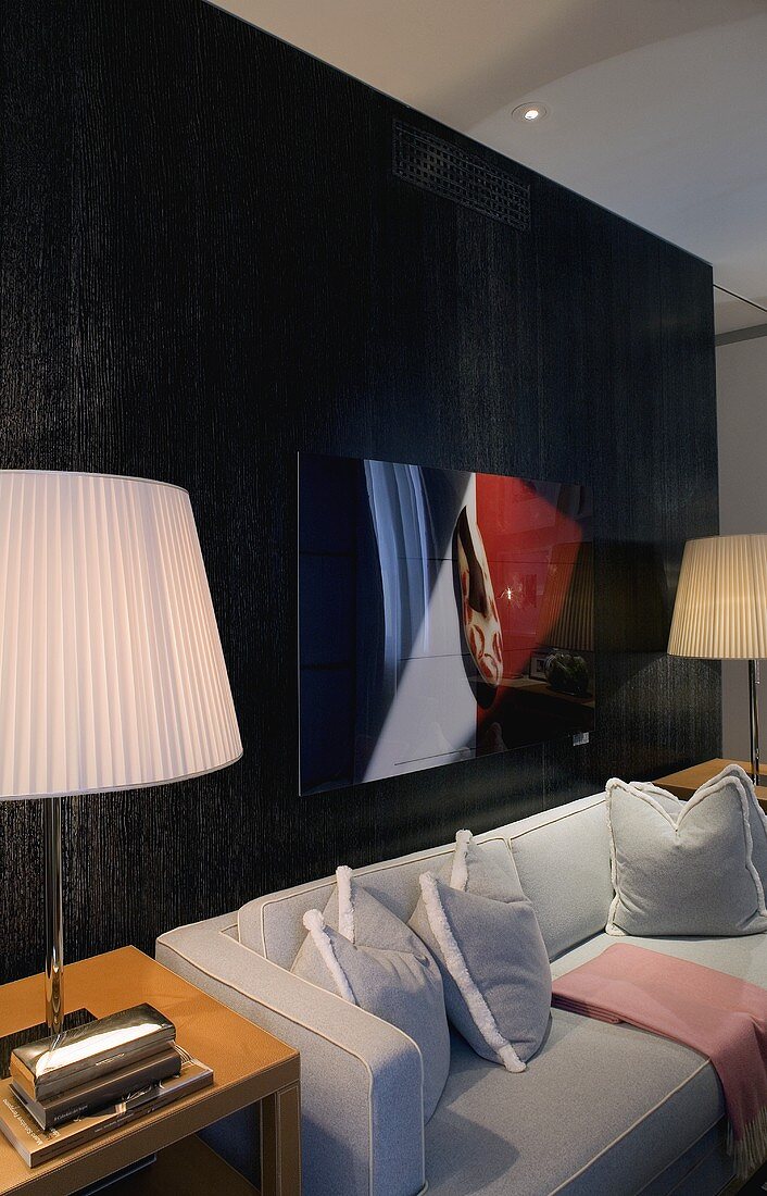 Hellgraues Sofa vor schwarz vertäfelter Wand mit Tischlampen und Plisseeschirmen