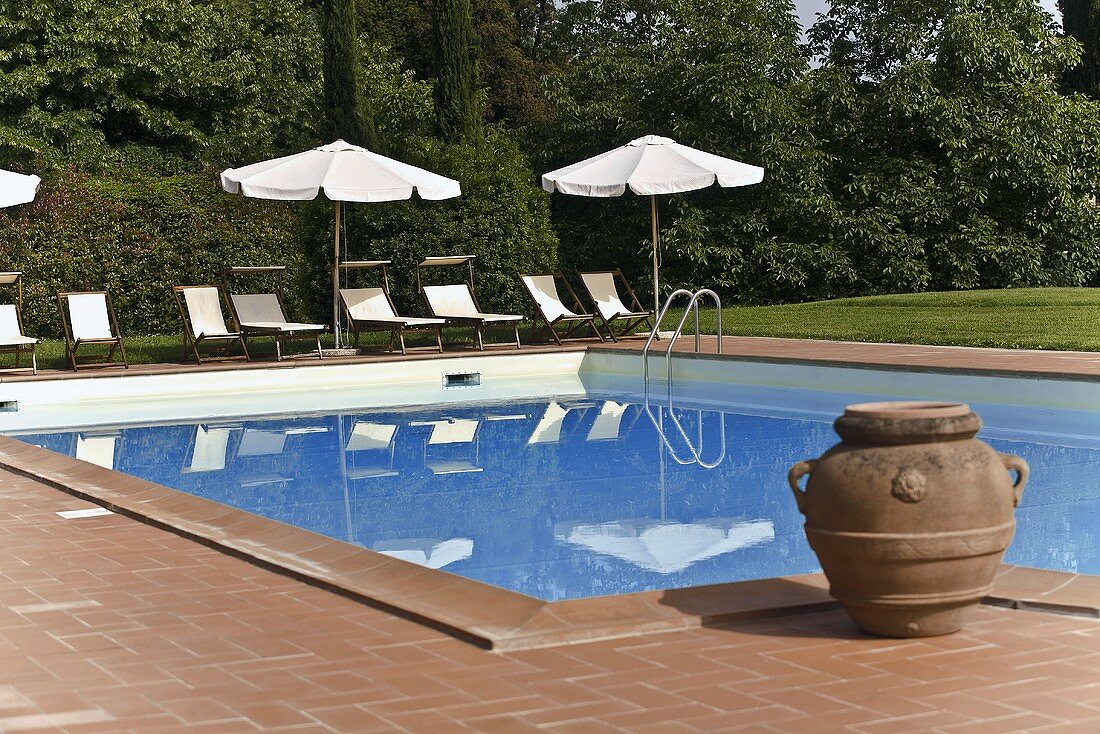Tongefäss am Pool und Liegestühle mit Sonnenschirm spiegeln sich im Wasser