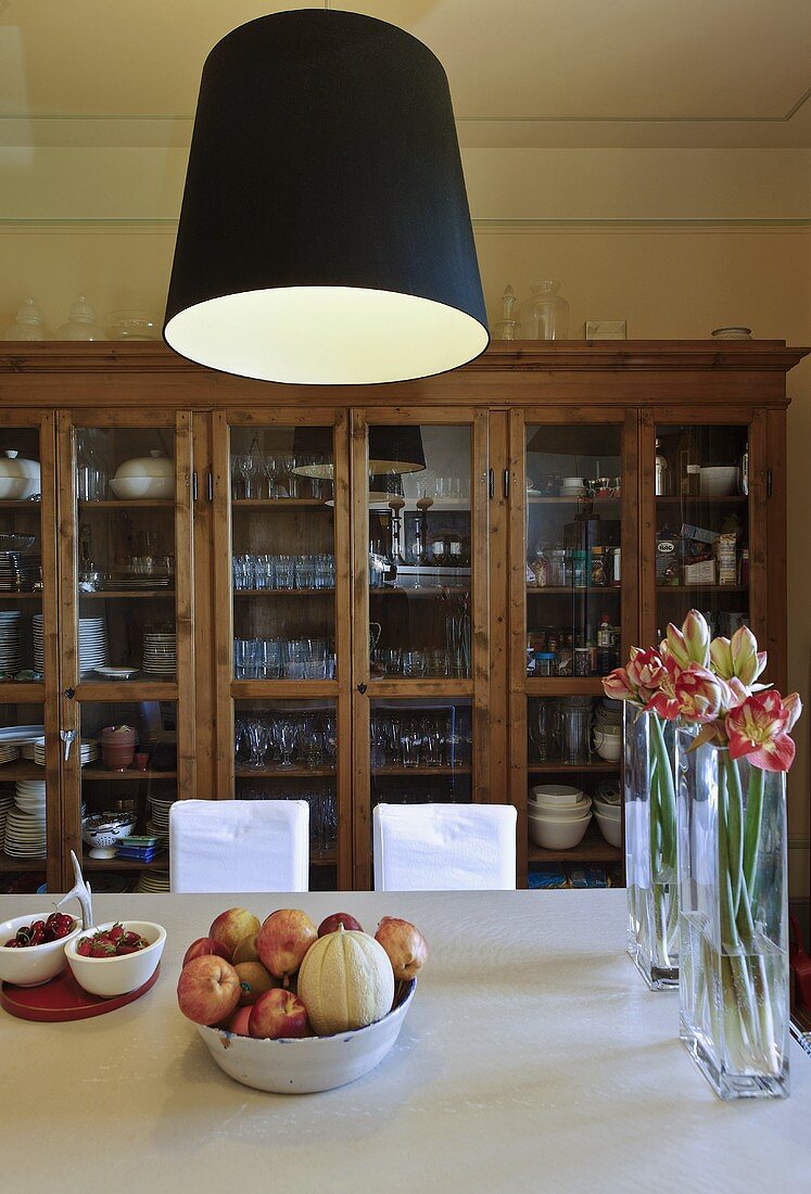 Black lampshades above a dining table with flower vases and a fruit bowl in front of a glass front cabinet