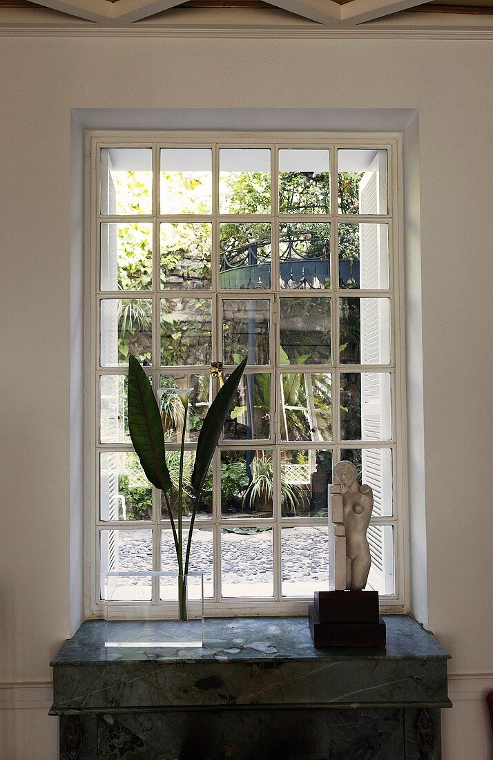 Palm fronds in a vase and stone statue in front of a mullioned window with a view of the garden