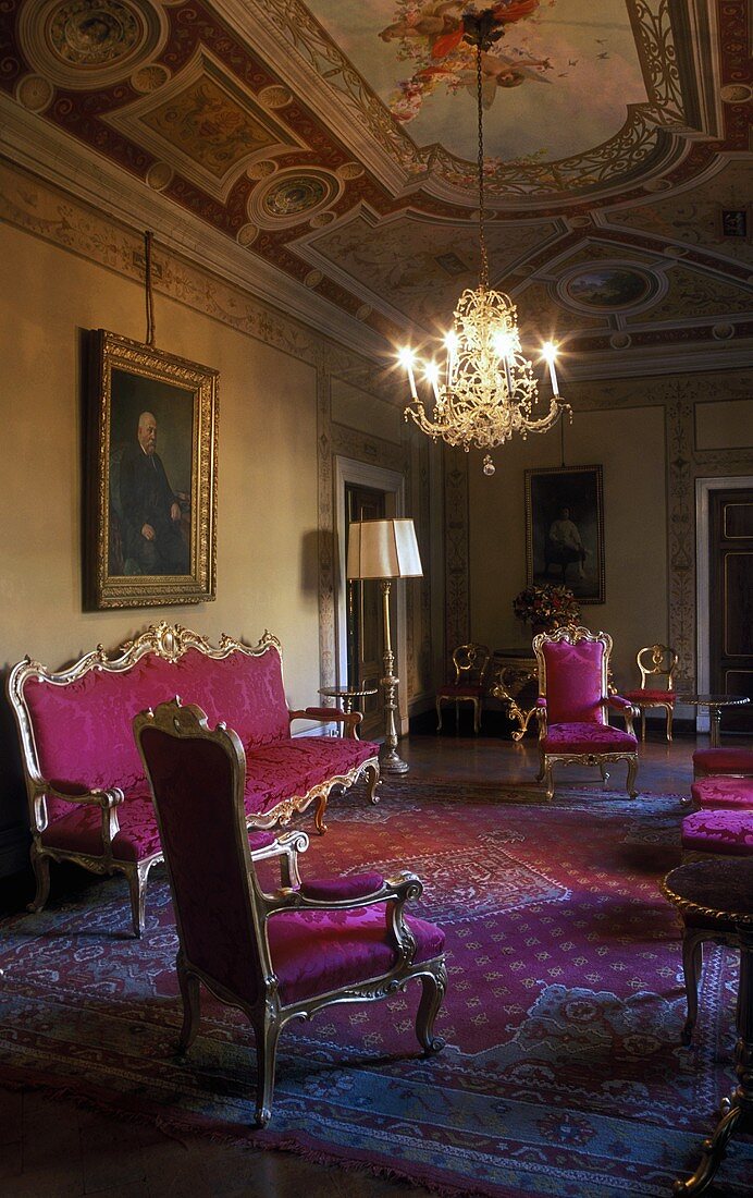 Rococo style sofa and chairs upholstered in red in the living room of a castle with a chandelier hanging from a frescoed ceiling