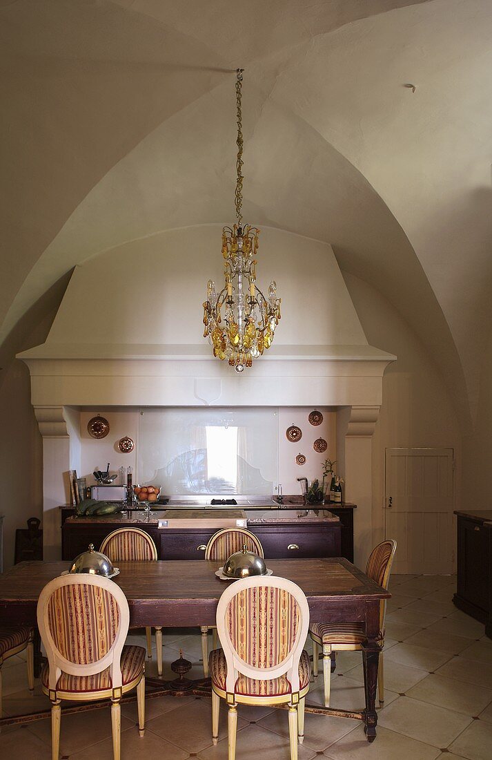 Esstisch mit gepolsterten Stühlen vor Küchenofen mit Kaminabzug und Kronleuchter unter Gewölbedecke