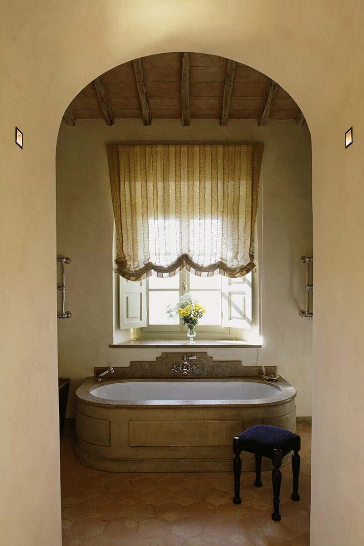 View through an archway of an elegant antique bathtub clad in stone and a stool upholstered in blue