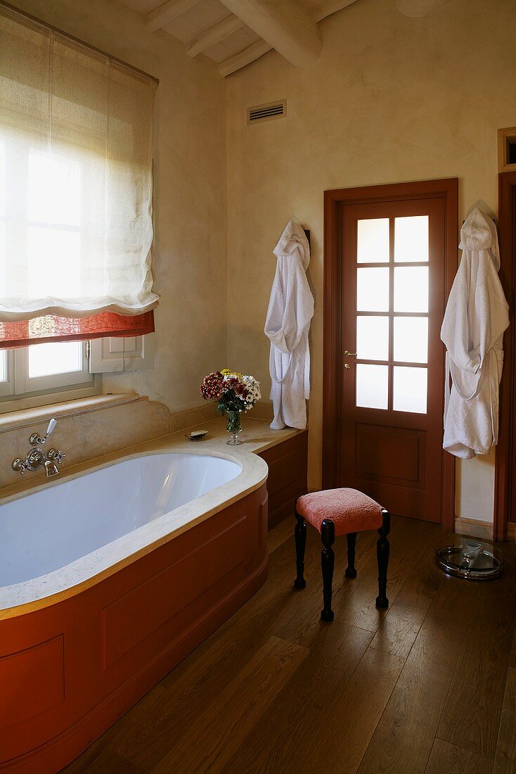 Country style bathroom -- bathtub clad in wood in front of a window with a Roman blind and upholstered stool on a wooden floor