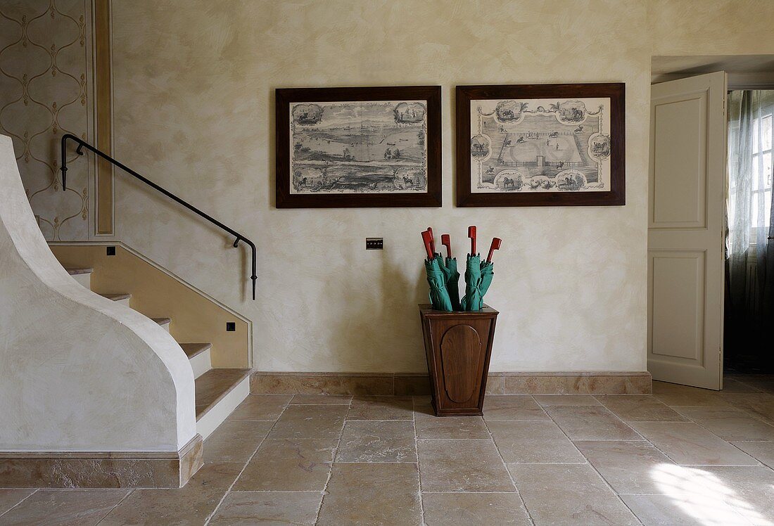 Cool foyer with stairs and umbrella stand in front of copperplate etchings on a wall and a stone floor