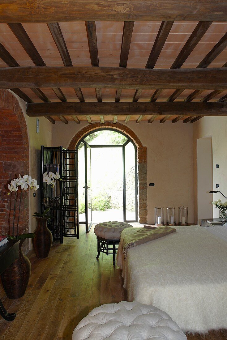 Elegant bedroom in a renovated country home with a rustic wooden beam ceiling and open terrace doors in a round arch