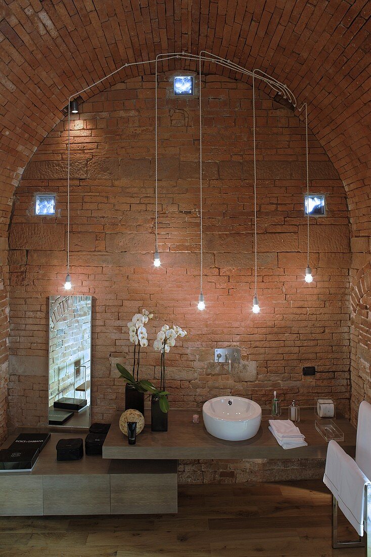 Waschtisch im Designerstil mit improvisierter Beleuchtung aus hängenden Glühbirnen vor Ziegelwand und Tonnendecke