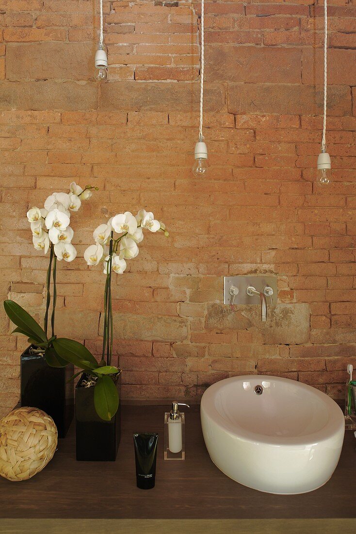 Waschtisch mit Keramikschüssel vor Ziegelwand und weiße Orchideen in Vase