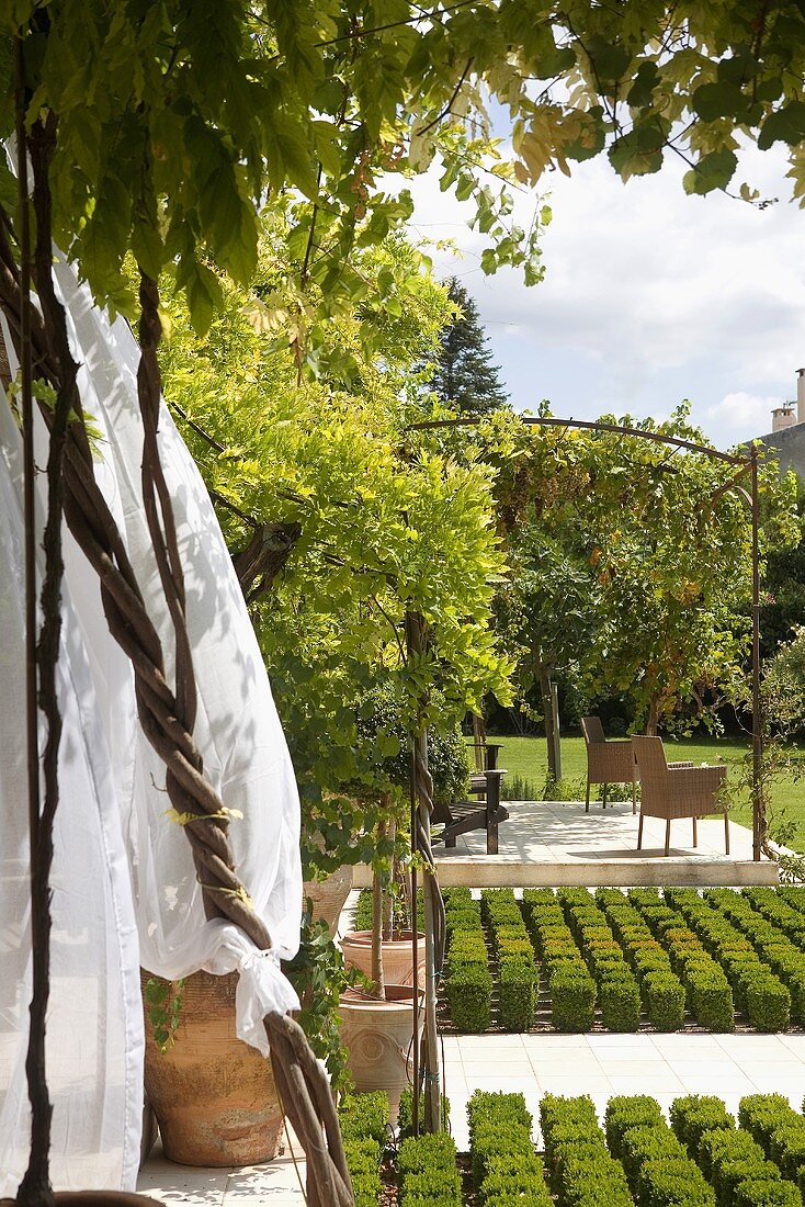 Blick über Rabatten mit Büschen in kubischer Form und Pergola mit Stühlen