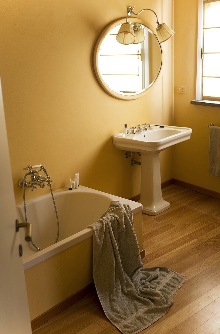 Country style bathroom - pedestal sink with a mirror in front of a yellow wall and gray bath towel draped over the bath tub