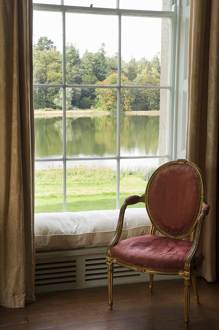 A pink padded chair with a gilded frame in front of a window with a view
