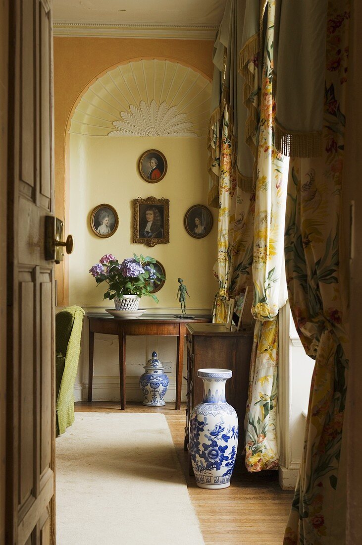 A view through an open door onto a blue and white floor vase and an occasional table in front of a yellow wall niche