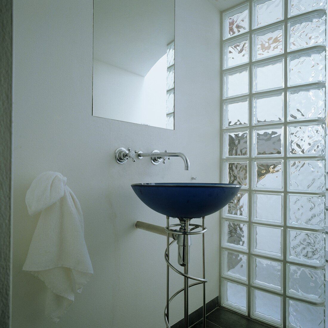A blue, free-standing wash basin in front of a glass brick wall and a wall tap under a mirror