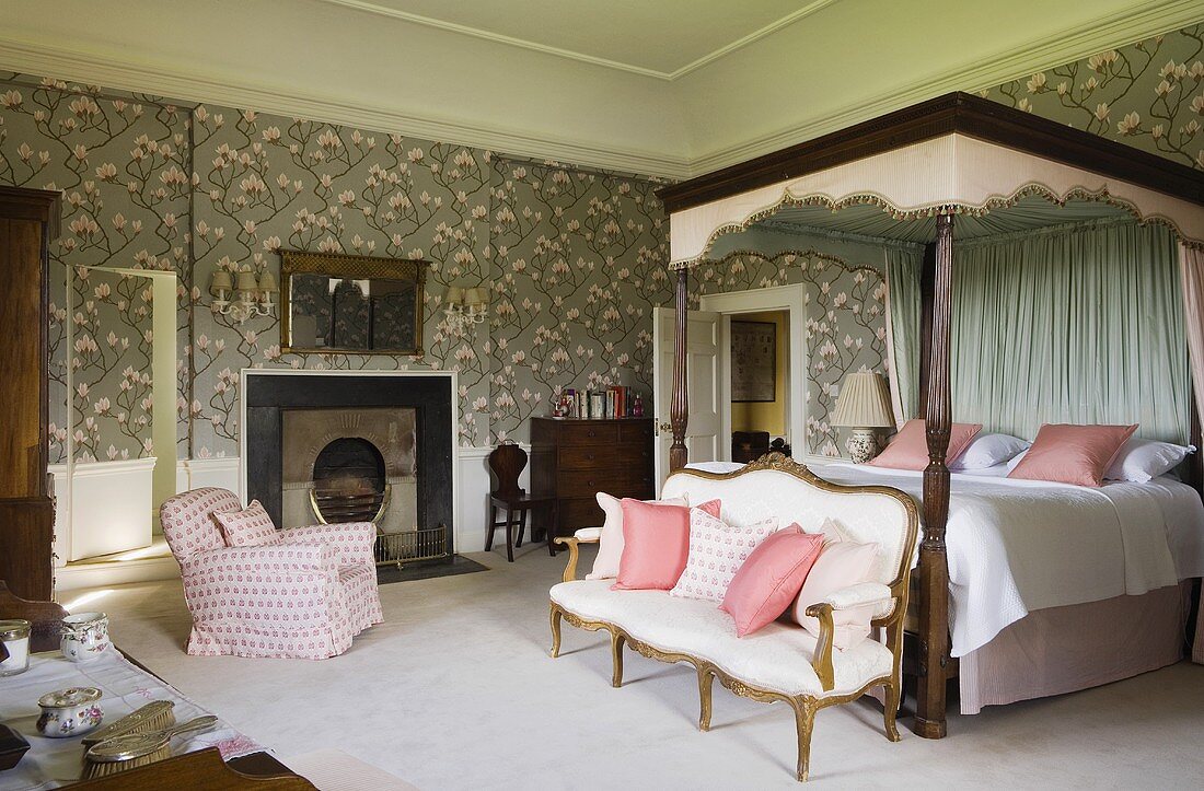 An elegant bedroom in a castle with an antique sofa at the foot of a four-poster bed with a canopy against a wall with floral wallpaper