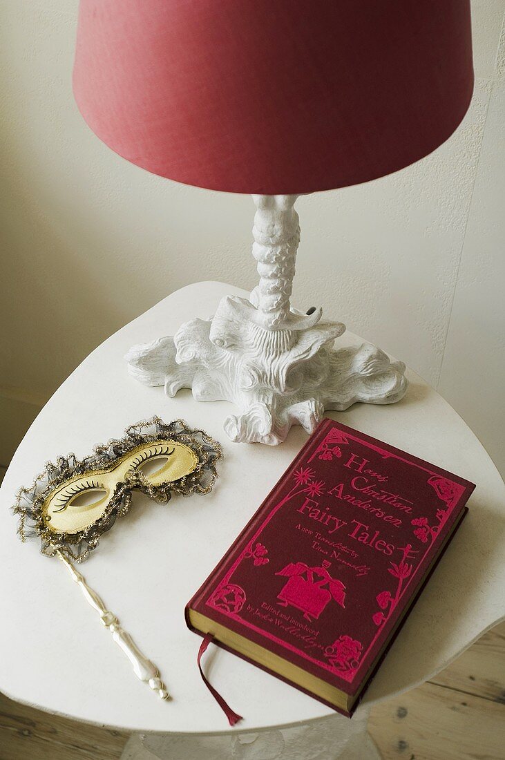Stillleben auf weissen Beistelltisch- Tischlampe mit rotem Schirm, venezianische Maske und ein Buch mit rotem Einband