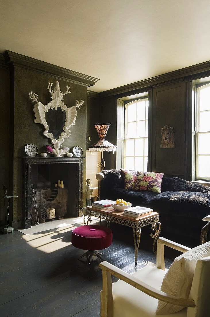 A dark painted fireplace room with green wooden panelling and a pink upholstered stool with an occasional table on the floor boards in front of the fireplace