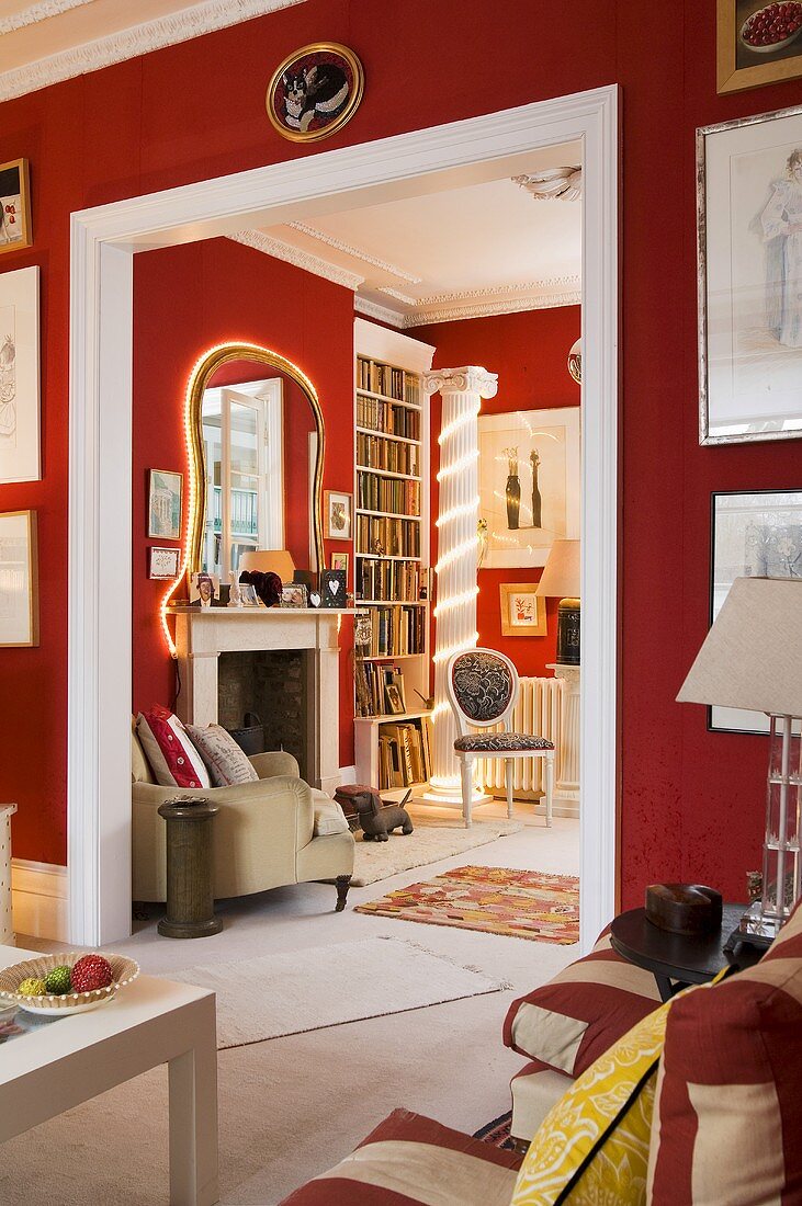 A living room with red walls and a white framed doorway with a view onto an illuminated mirror and a fireplace