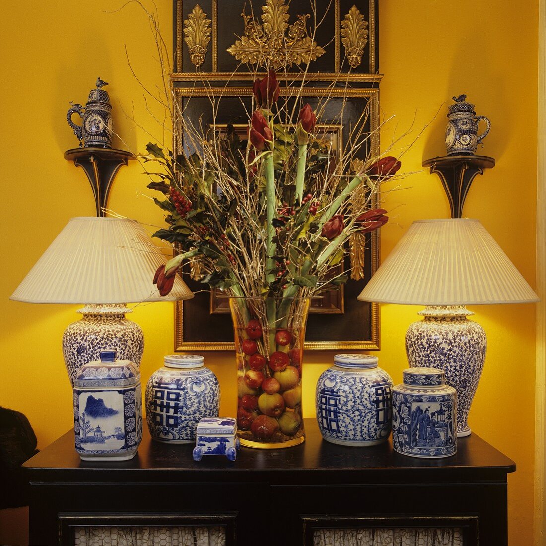 Chinese vases with plissé shades and porcelain on a chest of drawers with a bunch of amaryllis against a bight yellow wall