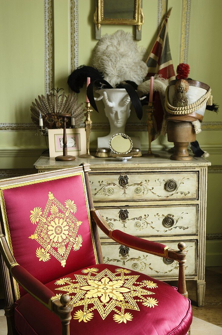 A baroque wooden chair upholstered in shiny red with a gold pattern in front of a painted chest of drawers