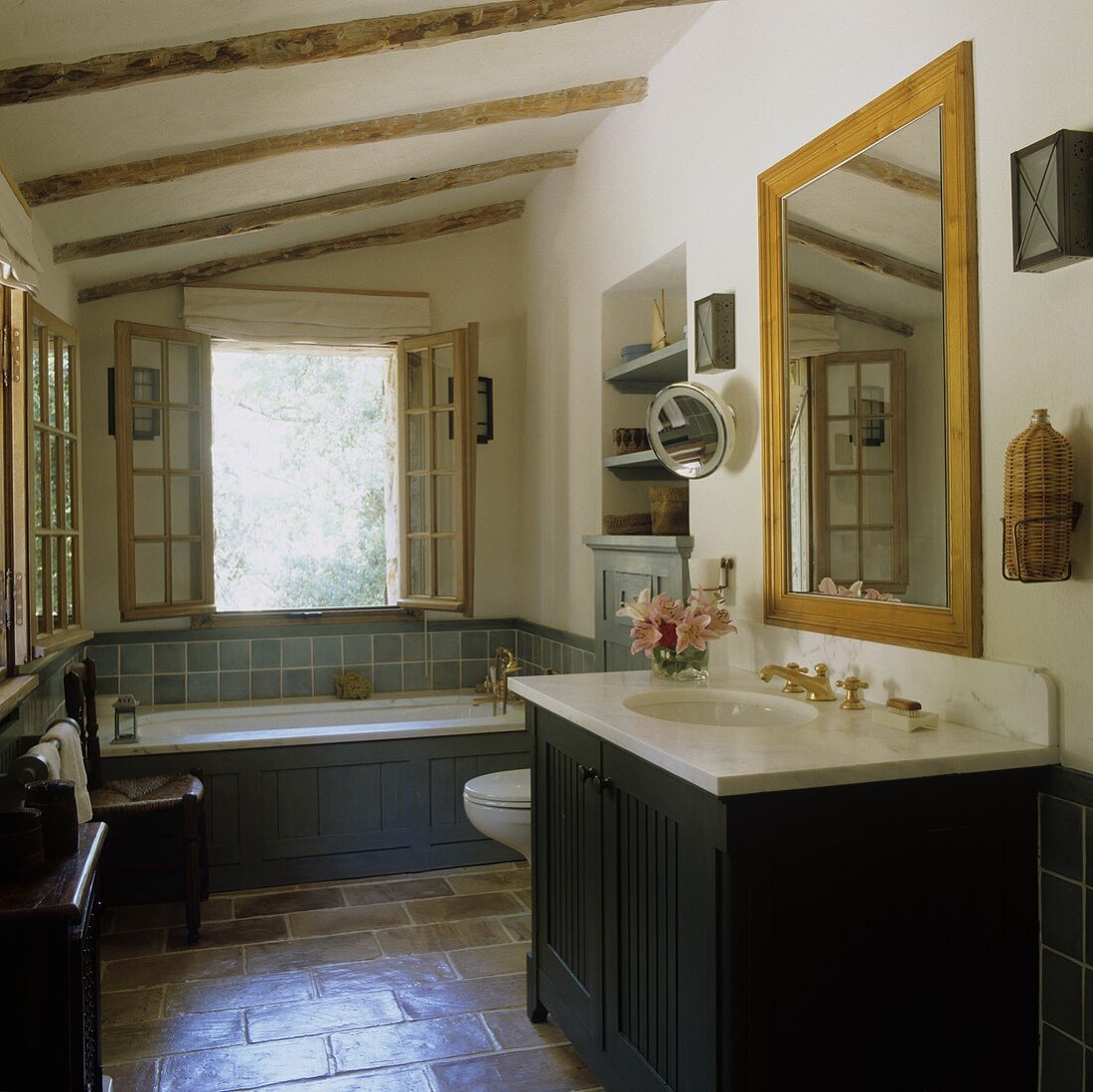 A bathroom in a country house with a terracotta floor - dark wood panelling on a wash basin and a bathtub in front of an open window