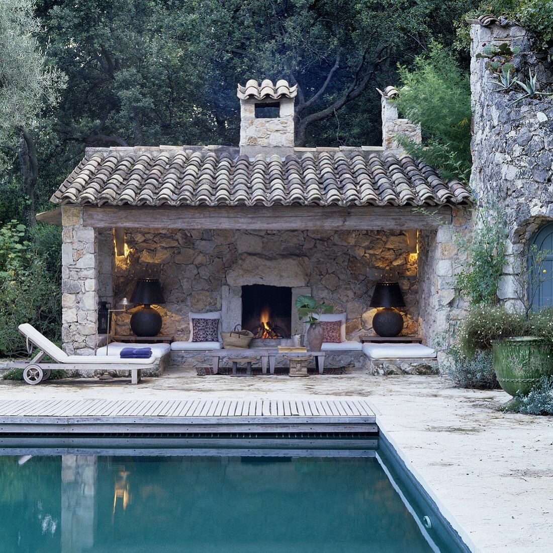 Pool und offener Anbau im Mediterraner Stil mit stimmungsvollem Licht vor Natursteinwand und Kamin
