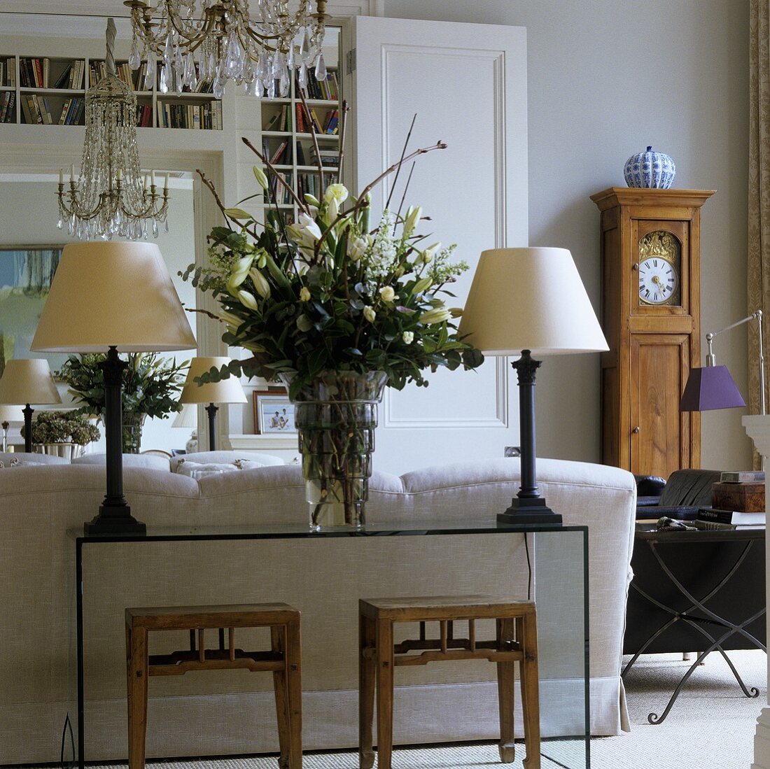 Table lamps with white shades and a bunch of lilies on a glass table with rustic stools and an antique grandfather clock
