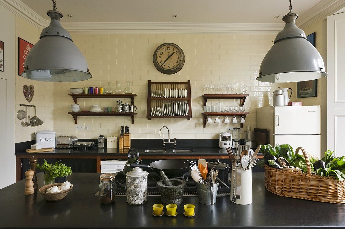 An open-plan kitchen in a country house - metal lamps hanging above a black work surface with kitchen utensils and a plate rack hanging on the wall