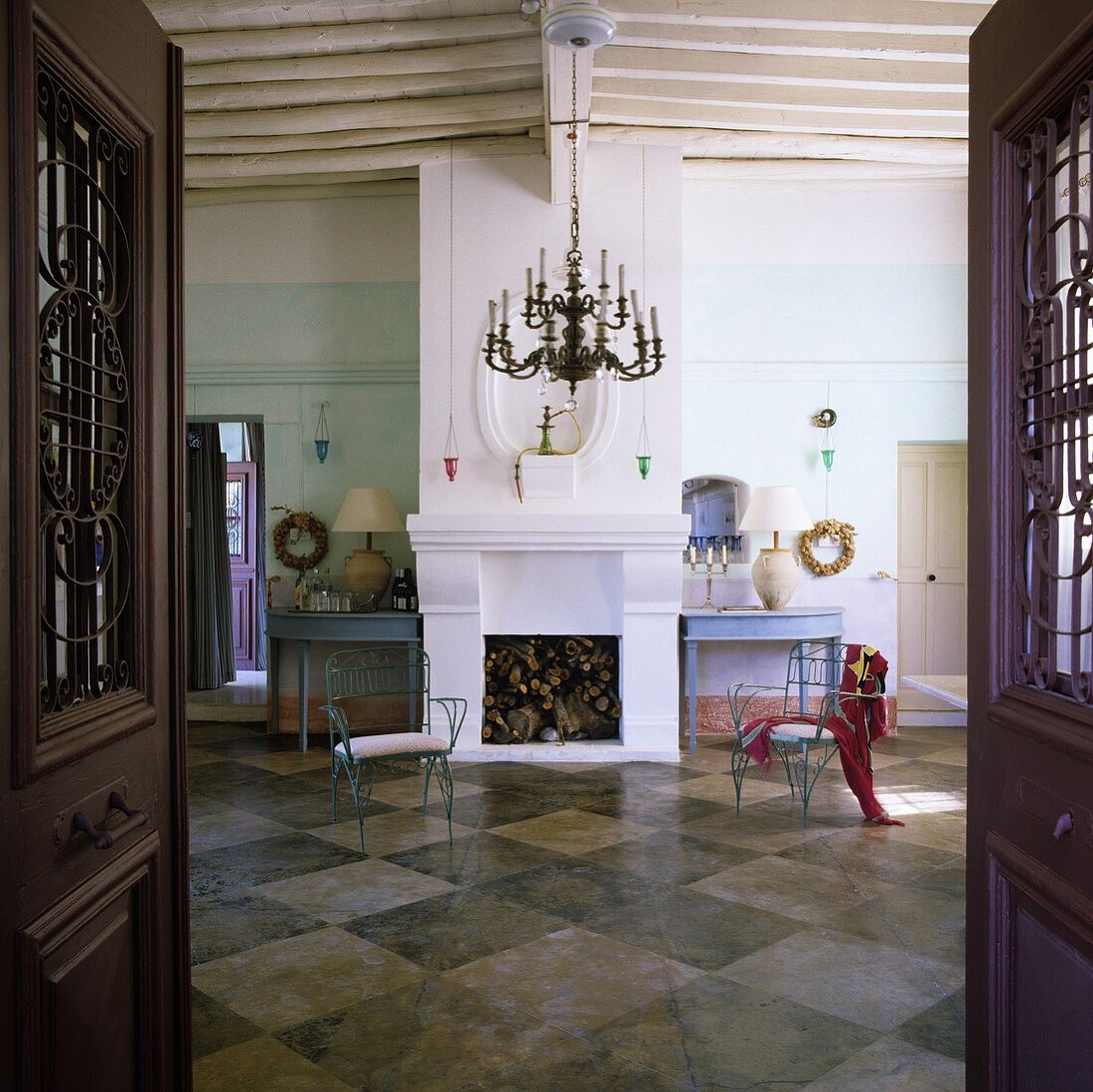 A view through an open door into a spacious living room with a chimney in a Mediterranean house and blue metal chairs on a black and white tiled floor