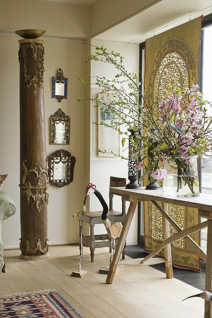 Ethnoflair im Wohnraum - antike Holzsäule und heller Holztisch vor folkloristischem Tuch am Fenster