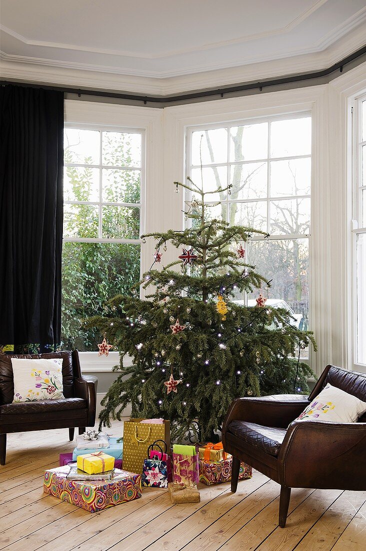 A leather armchair next to a decorated Christmas tree with presents in the bay window of a living room