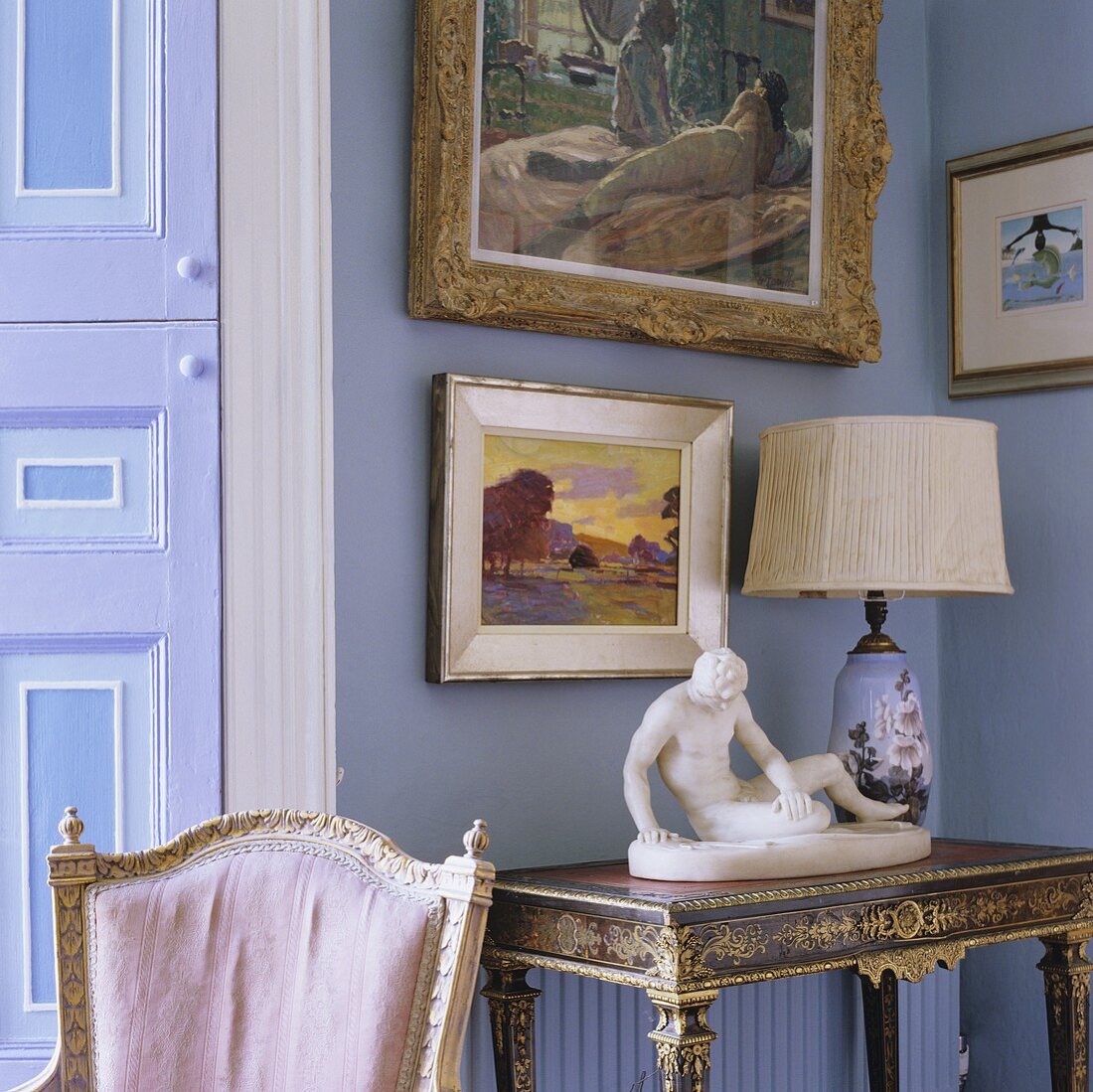 A sculpture and a table lamp with a white shade on an antique wall table and pictures hanging on a lilac-painted wall