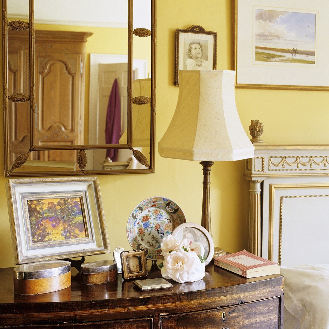 A table lamp with a white shade on a bedside table and a mirror above it on a yellow wall