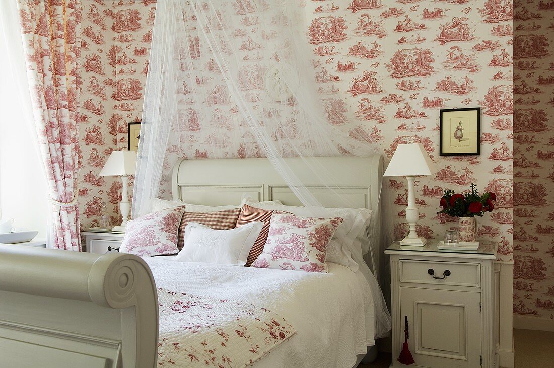 A white wooden country house-style bed with pillows, curtains and matching wallpaper