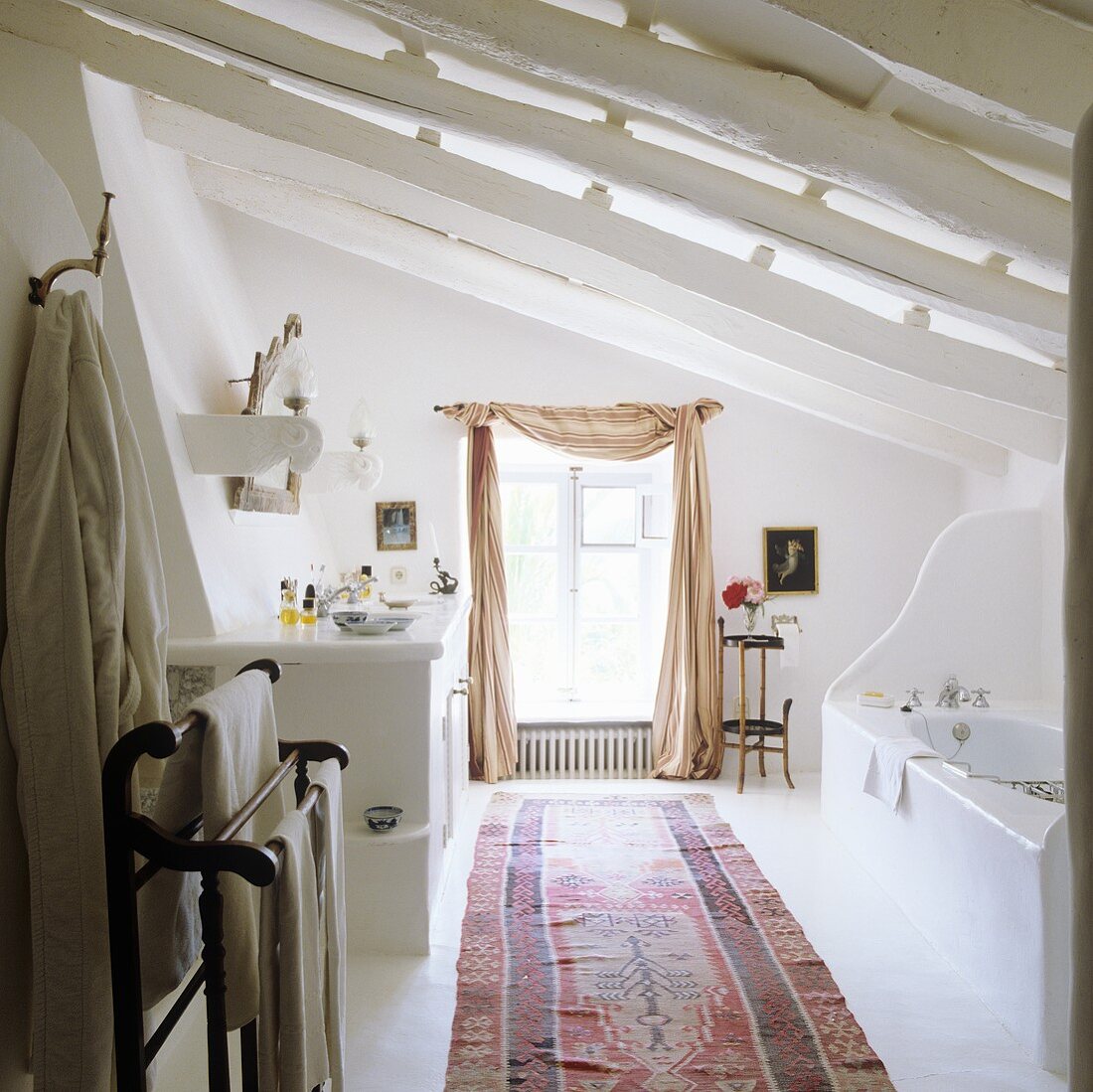 Mediterraner Bad unter dem Dach mit weiss gestrichenen Holzbalken und Teppichläufer vor Fenster mit drapiertem Vorhangschal