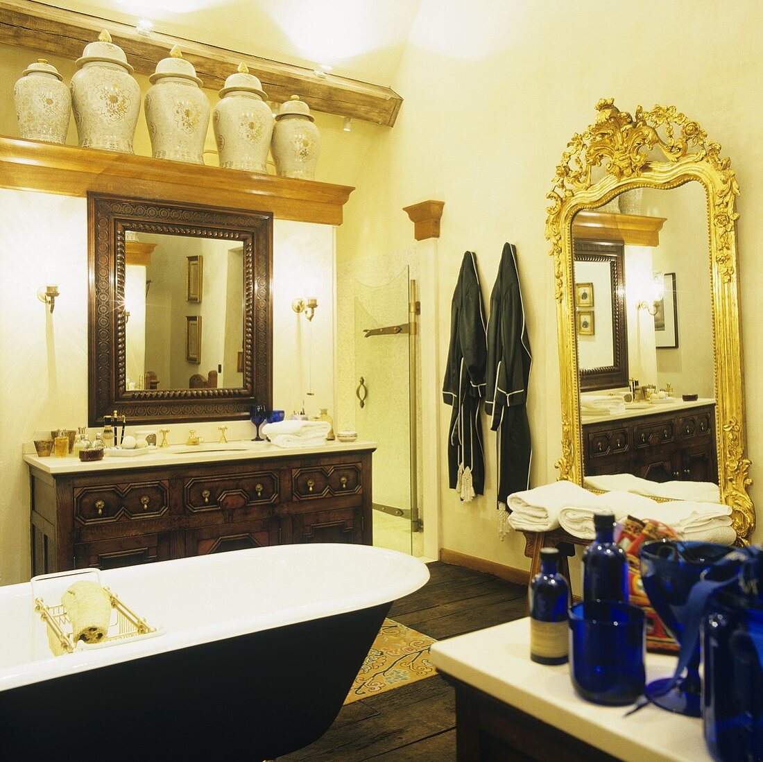 A bathroom in a country house with a free standing bathtub in front of a wash basin with a wooden cupboard and a wall mirror with a golden frame