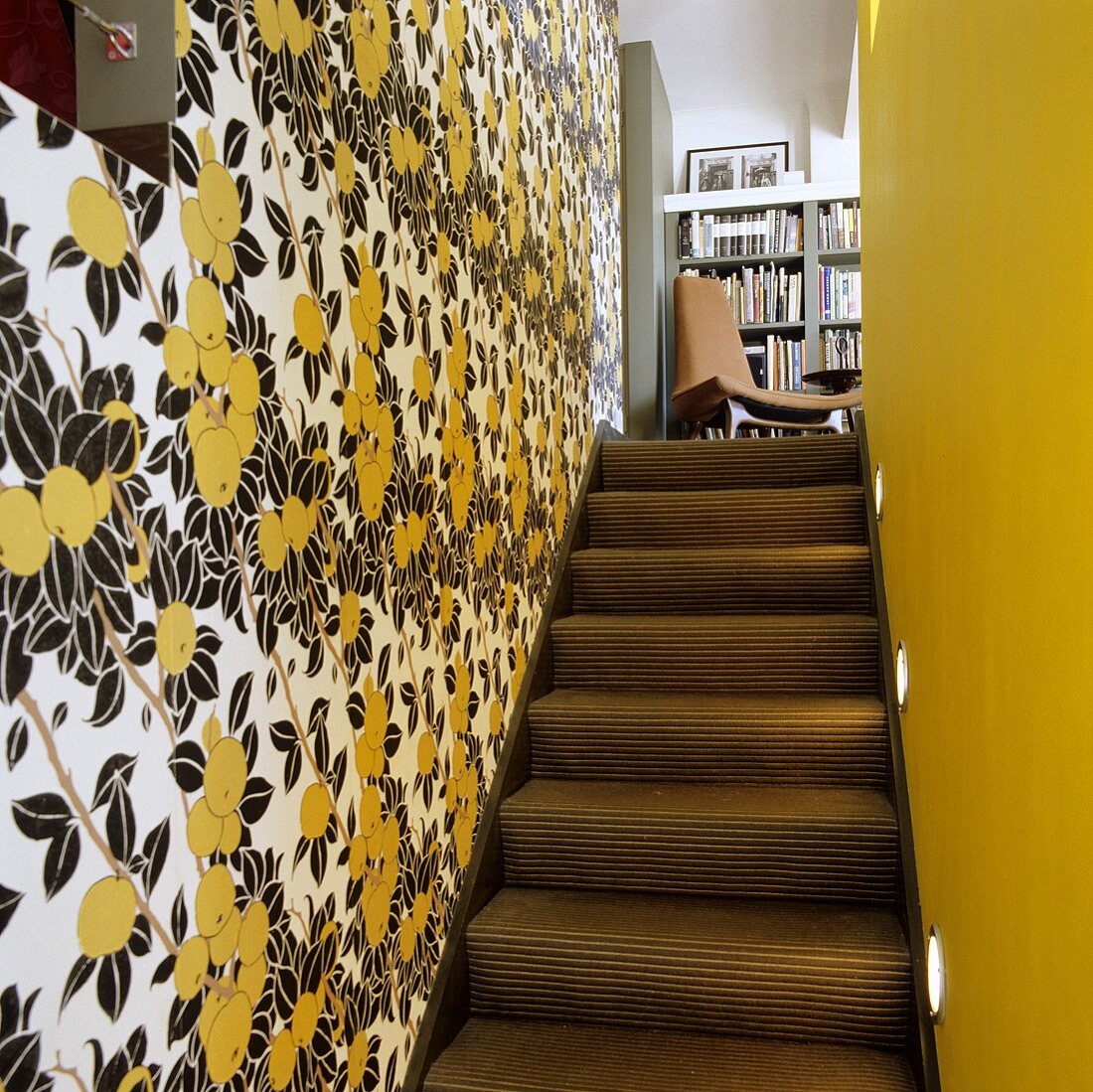 A narrow stairway with a flower-patterned papered wall and a yellow-painted wall with a view onto a chair