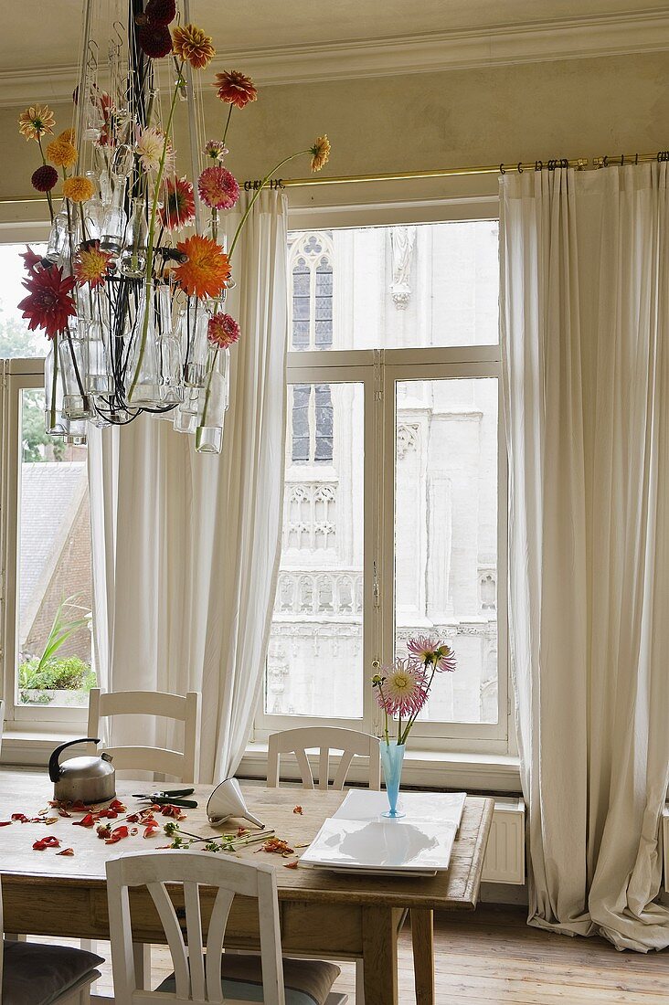 A dining area in front of a window with white floor-length curtains and glasses hanging from the ceiling filled with flowers