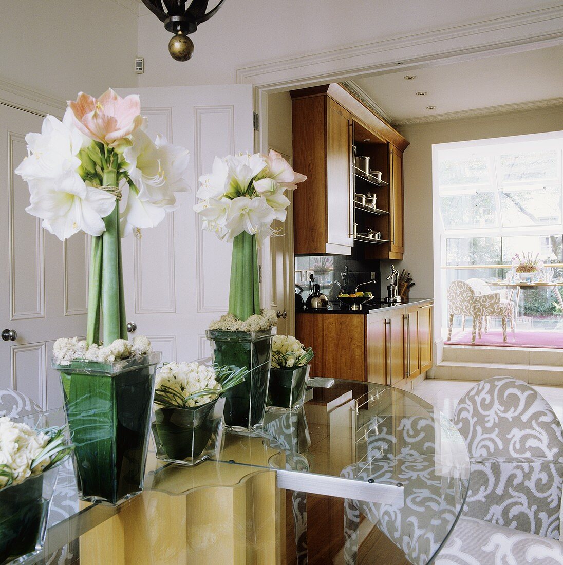 White amaryllis in a glass vase on a glass table with open swing doors and view of a fitted kitchen