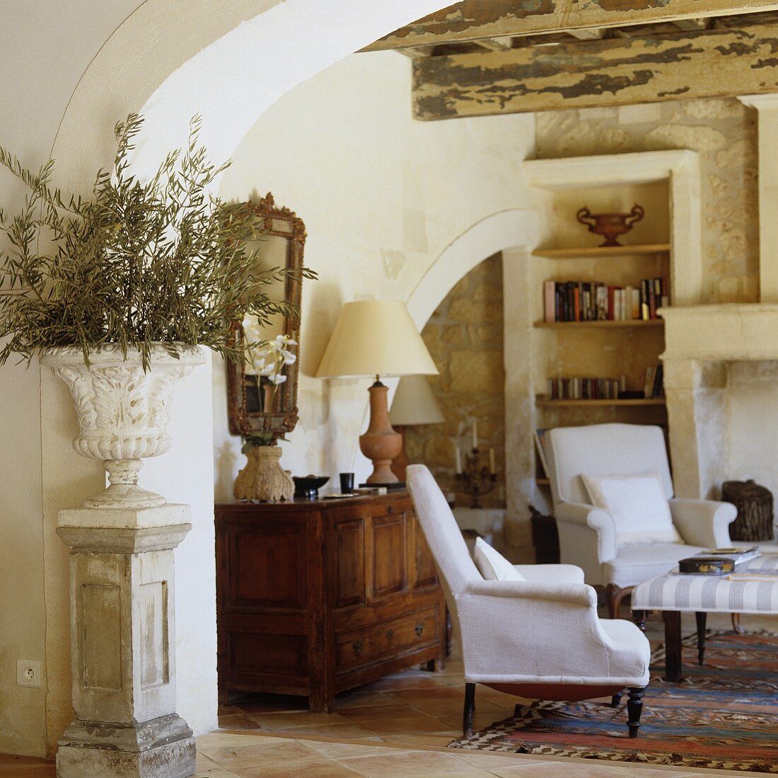 A view through a round archway into a corner of a rustic living room with white armchairs in front of a wooden sideboard