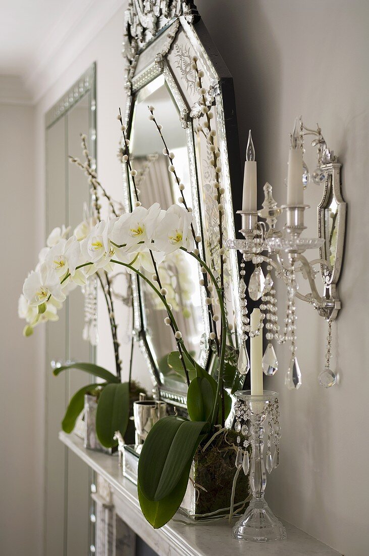 weiße Orchideen auf Kaminsims und Wandkerzenleuchter mit Kristallschmuck neben Spiegel