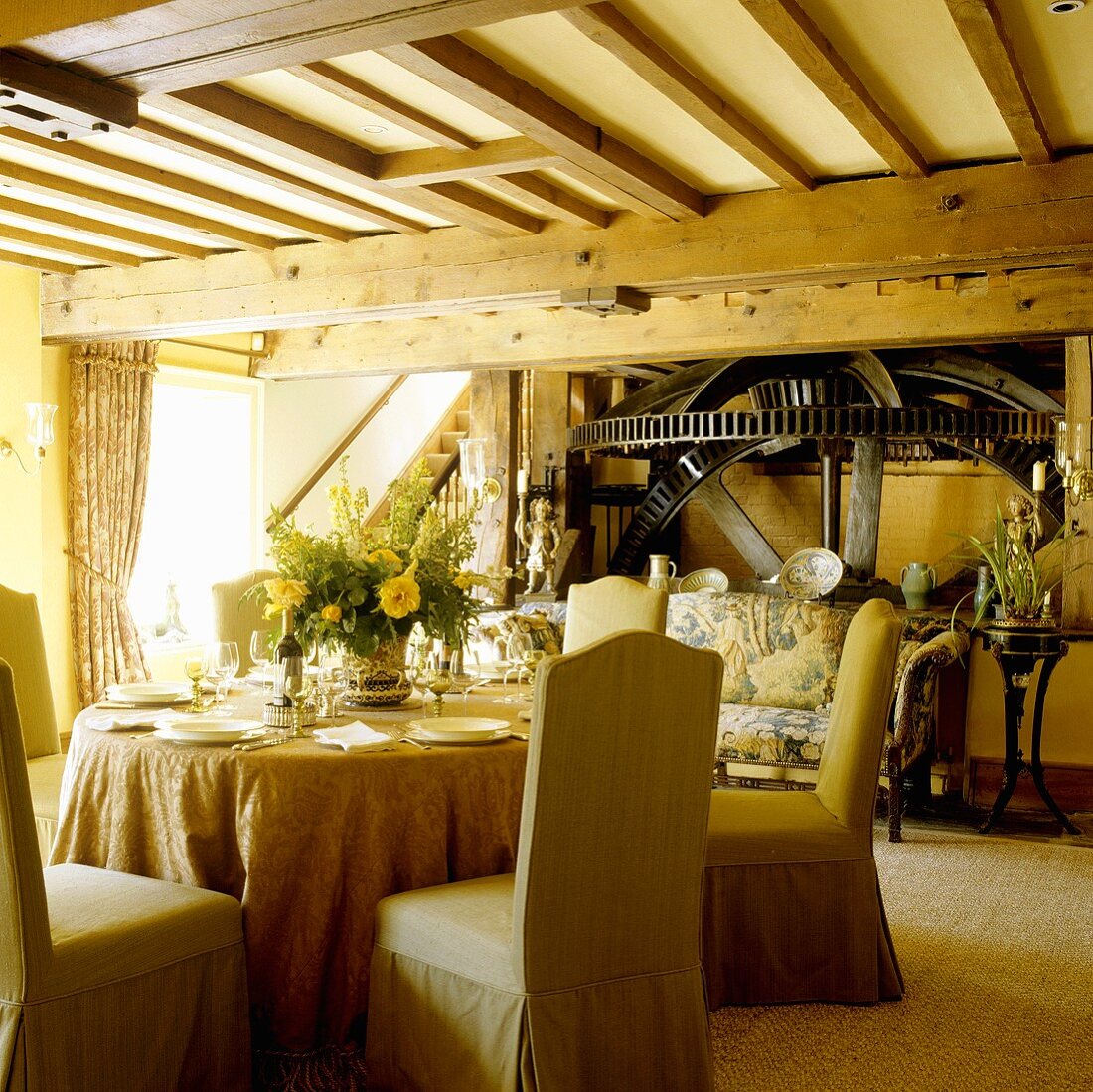 An old English mill house - covered chairs around a laid table in an open-plan living room with a wood beam ceiling