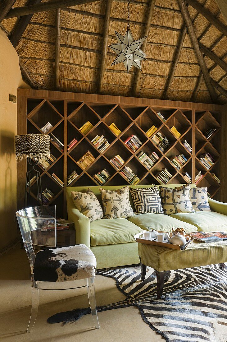 Hellgrünes Sofa und Acrylstuhl mit Fellbezug vor Holzbücherregal in Wohnraum in südafrikanischen Villa