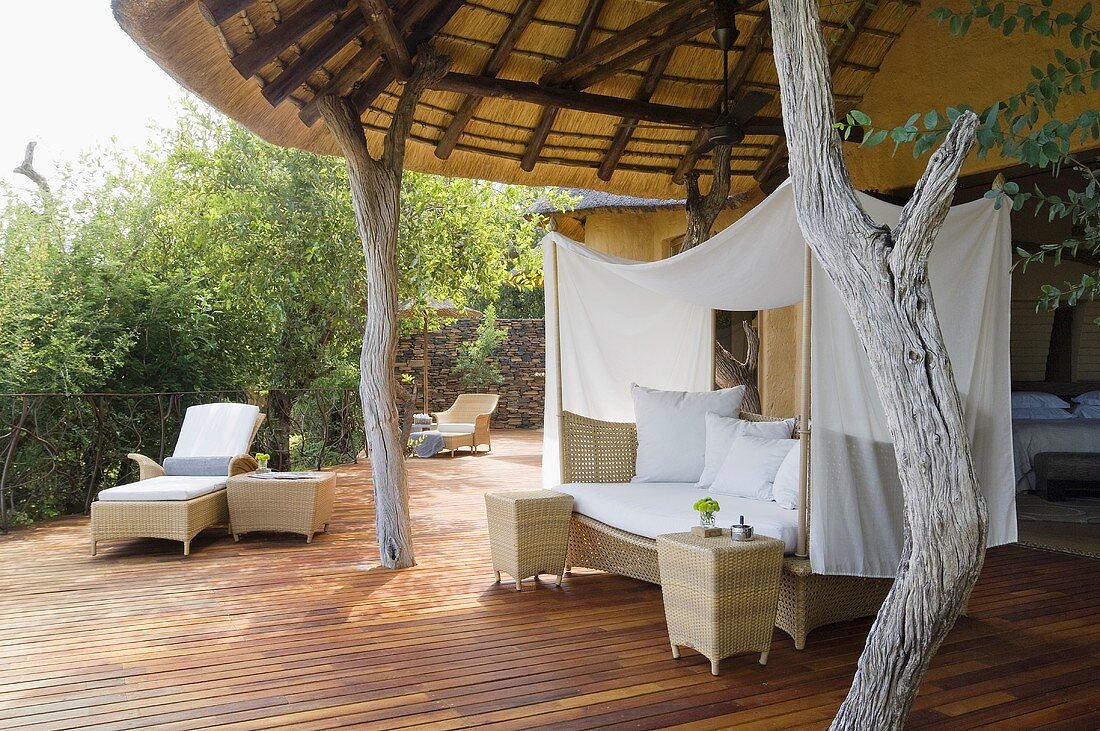 Korbsofa mit weißem Baldachin und rustikalen Holzstützen auf Terrasse eines südafrikanischen Hauses
