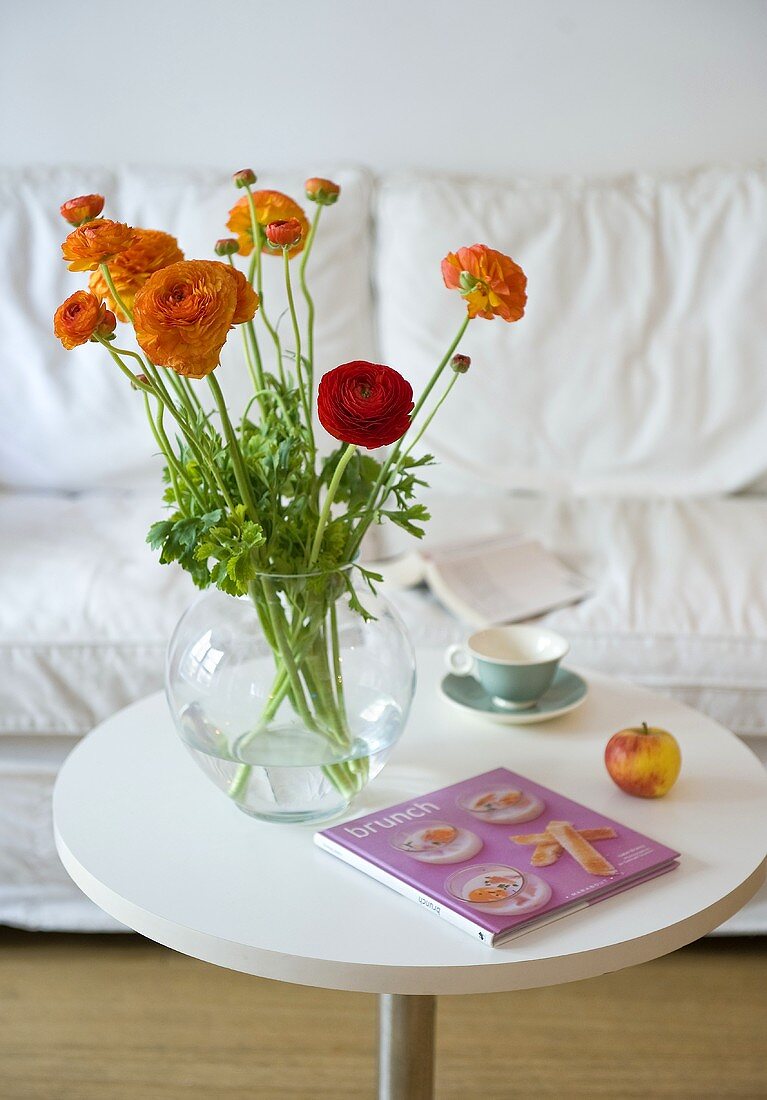 Orangefarbene Blumen in Glasvase auf weißem Bistrotisch vor Polstersofa