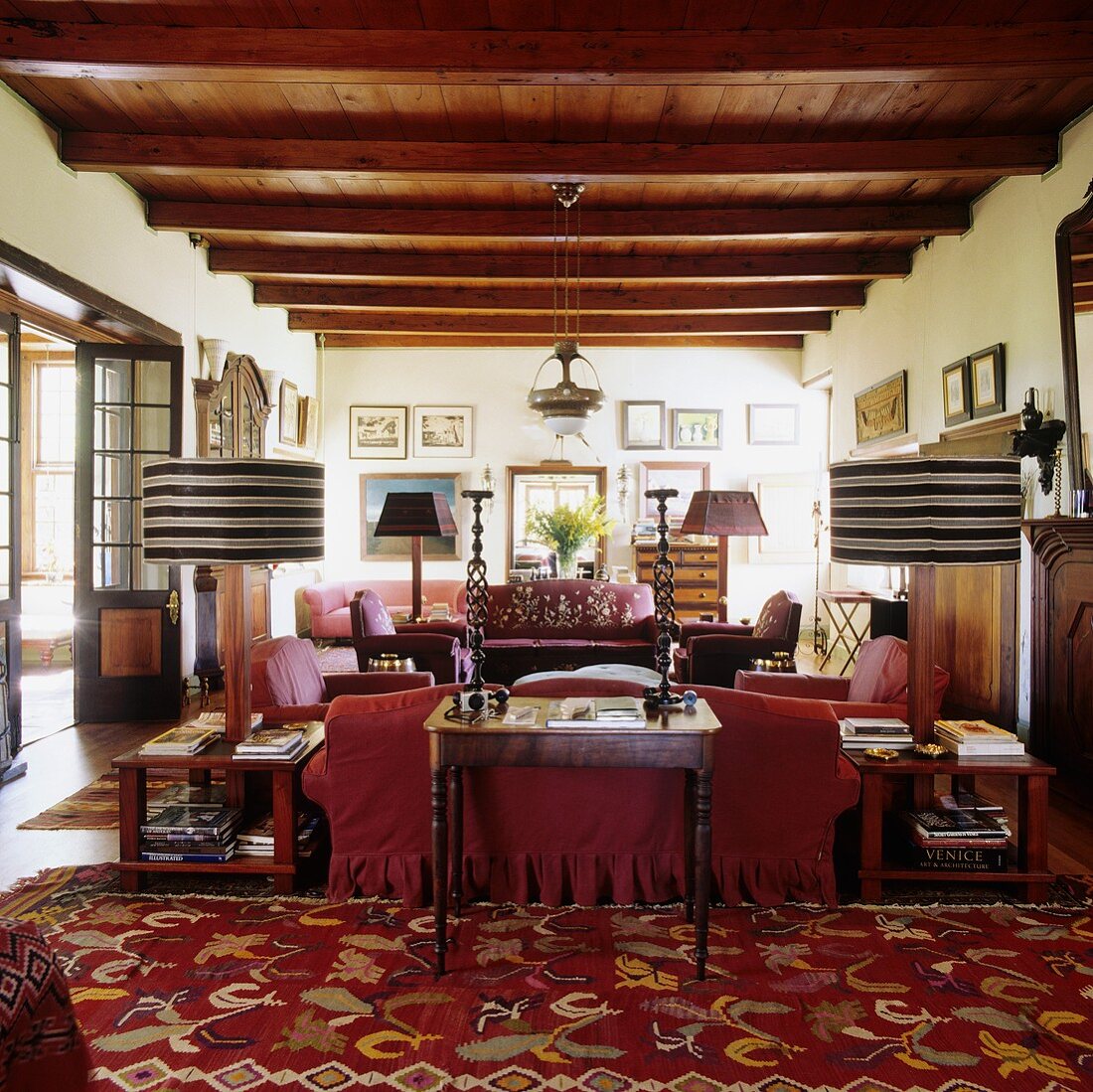 Wohnraum mit rustikaler Holzdecke im südafrikanischen Landhaus und roter Sofagarnitur mit Tischlampen und gestreiftem Schirm