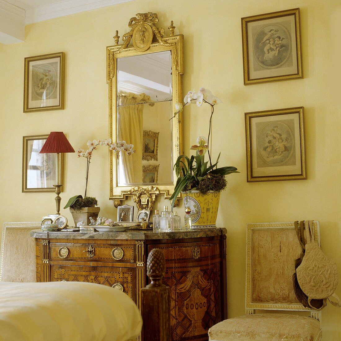 An antique wooden chest of drawers with a gold framed mirror on a yellow-painted wall in a bedroom